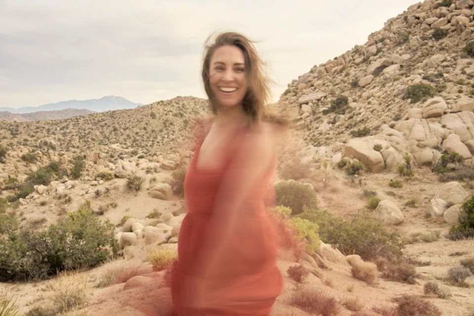 Smiling woman in a dress stands in a desert landscape with rocky hills under a cloudy sky, creating a blurred, dreamy photograph.