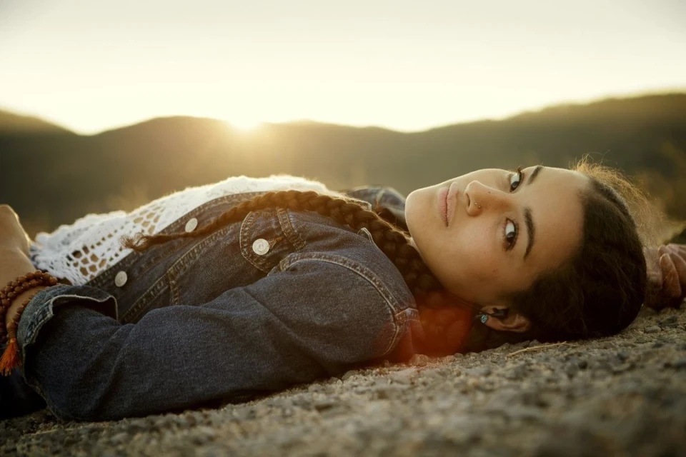 A young woman with braided hair and a denim jacket rests on a rocky surface with mountains and the sun setting in the background.