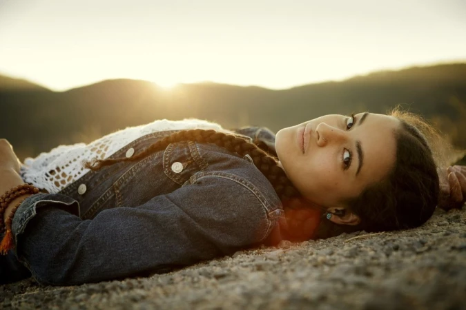 A young woman with braided hair and a denim jacket rests on a rocky surface with mountains and the sun setting in the background.