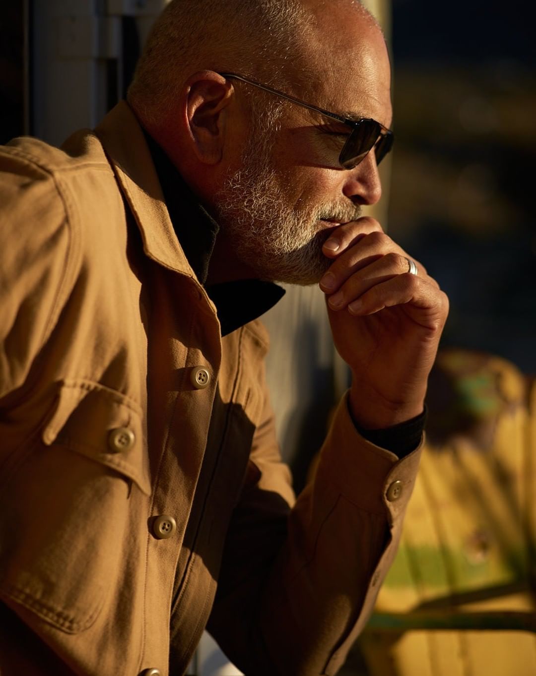Older man with sunglasses in a tan jacket sits with hand to face in contemplation, medium shot in a neutral setting with warm lighting.