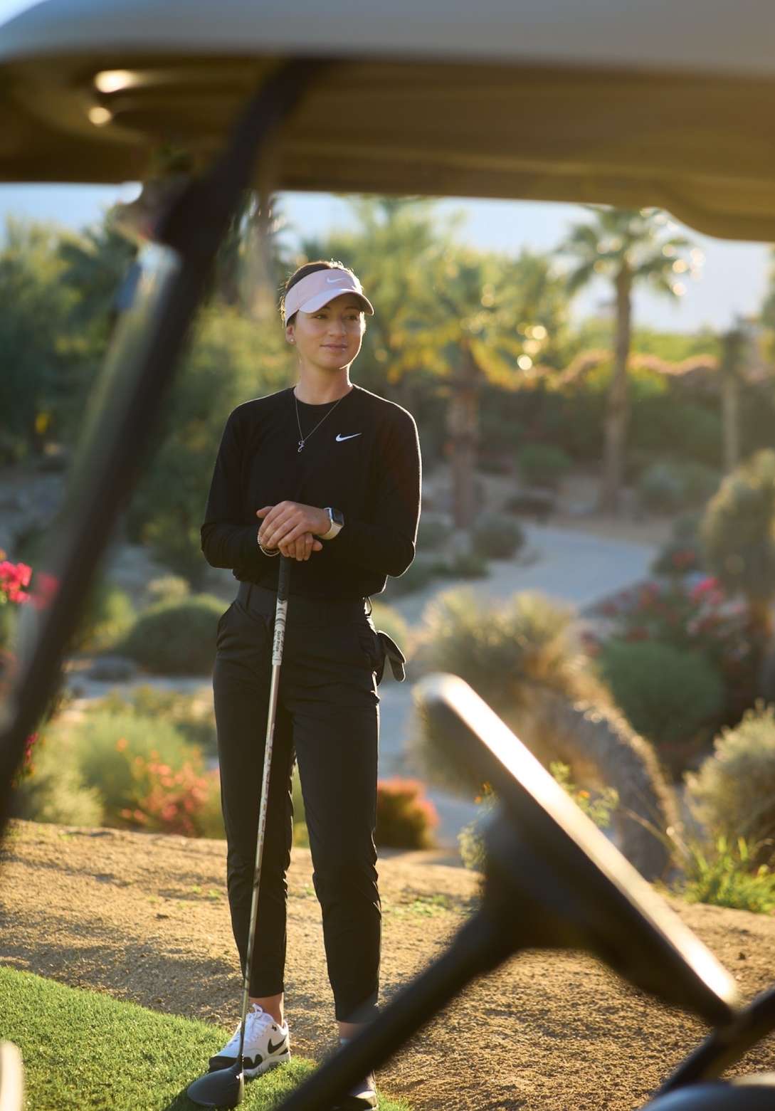 Female golfer in black sportswear stands on a green, sunny golf course with a golf club in hand, framed by a golf cart.