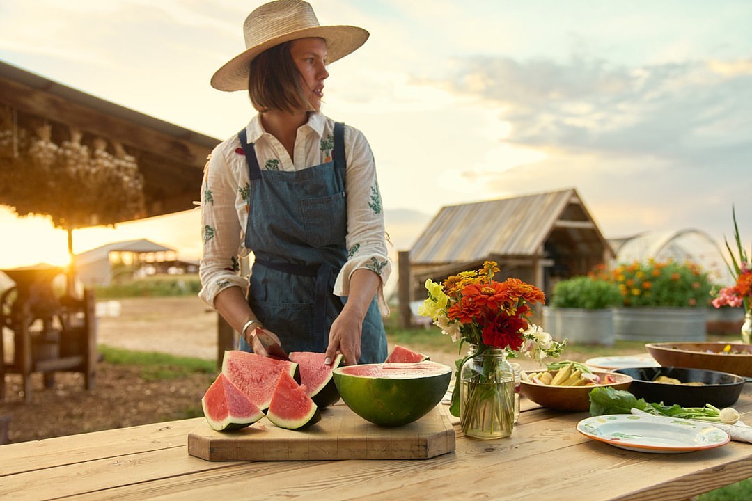 A woman in a hat and apron cuts watermelon at a table outdoors, with flowers, dishes, and farm buildings in the background at sunset.