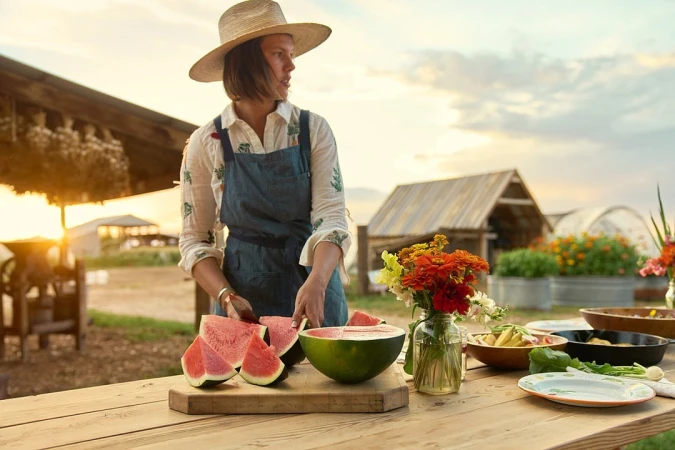 A woman in a hat and apron cuts watermelon at a table outdoors, with flowers, dishes, and farm buildings in the background at sunset.