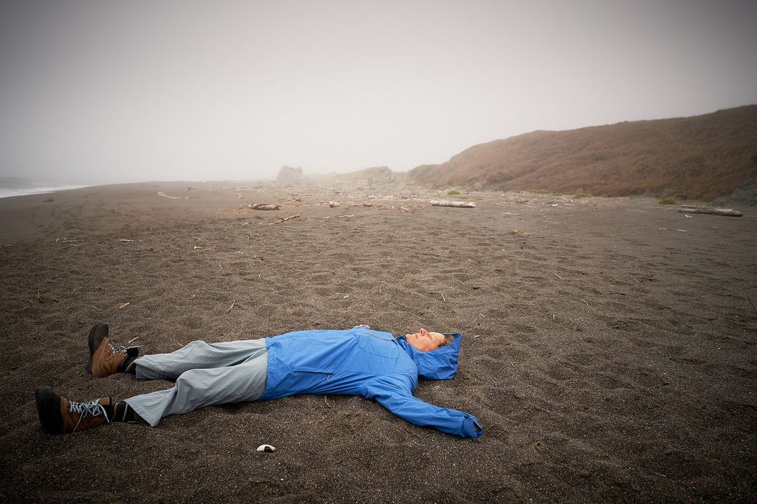 Person lies on black sand beach in blue hooded jacket and gray pants, wearing hiking boots; foggy coastal backdrop.