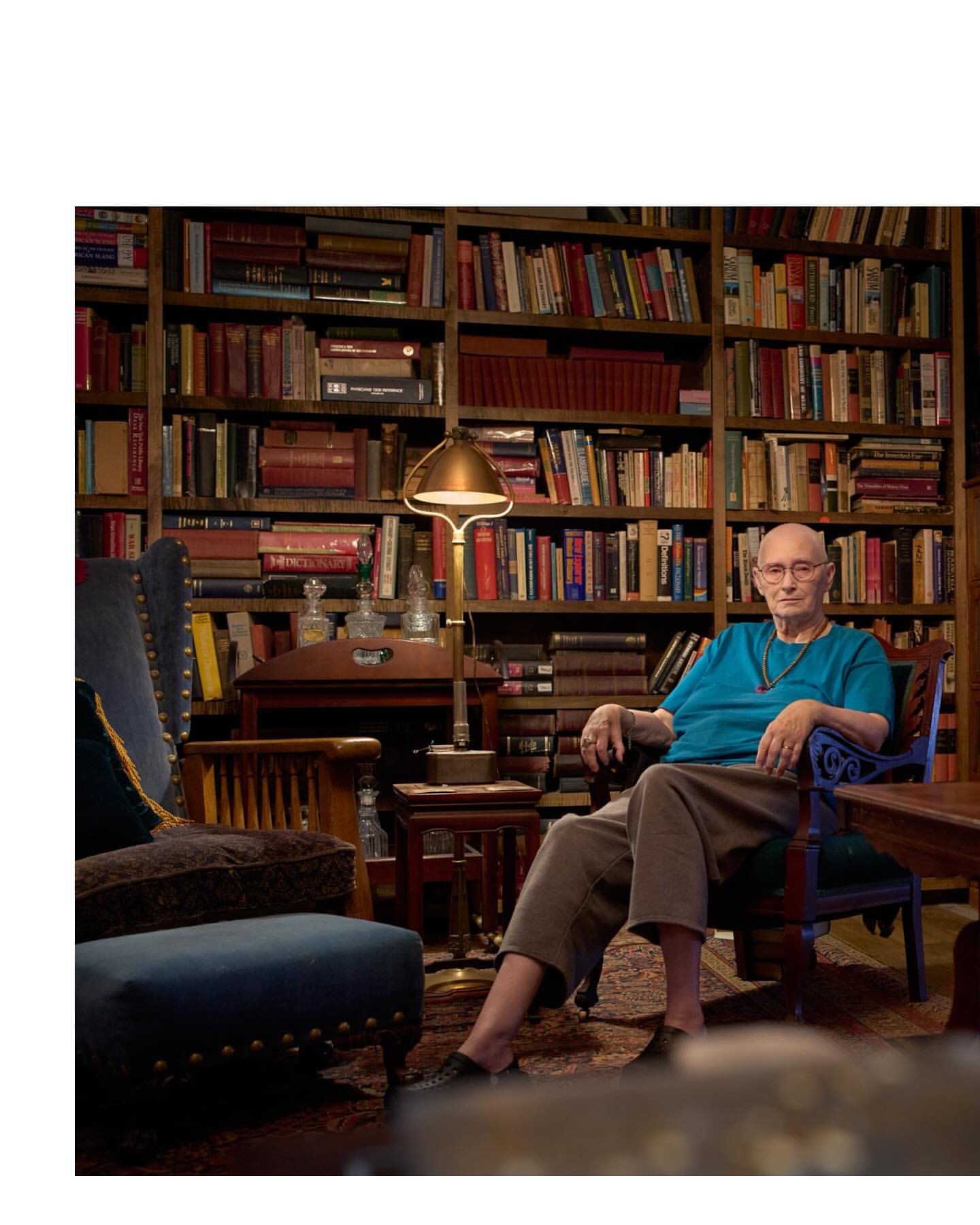 Elderly adult seated in blue armchair indoors, surrounded by a large bookshelf filled with books and illuminated by a floor lamp.