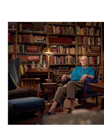 Elderly adult seated in blue armchair indoors, surrounded by a large bookshelf filled with books and illuminated by a floor lamp.