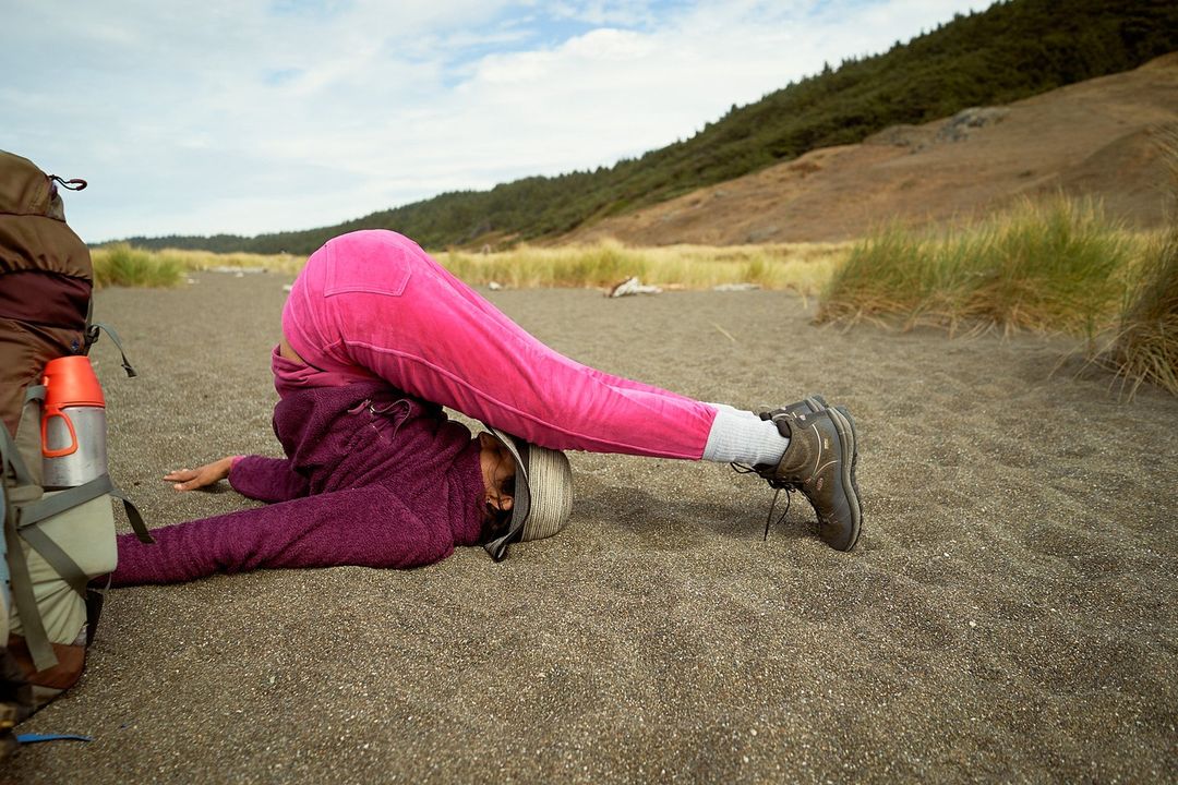 Adult female lying on a sandy beach, legs raised in the air, wearing a purple jacket, pink pants, and hiking boots, outdoors.