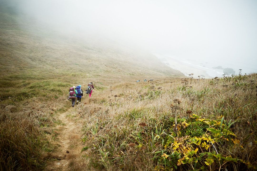 Hikers with backpacks ascend a grassy trail on a foggy hillside, showcasing an outdoor adventure and challenging terrain with limited visibility.
