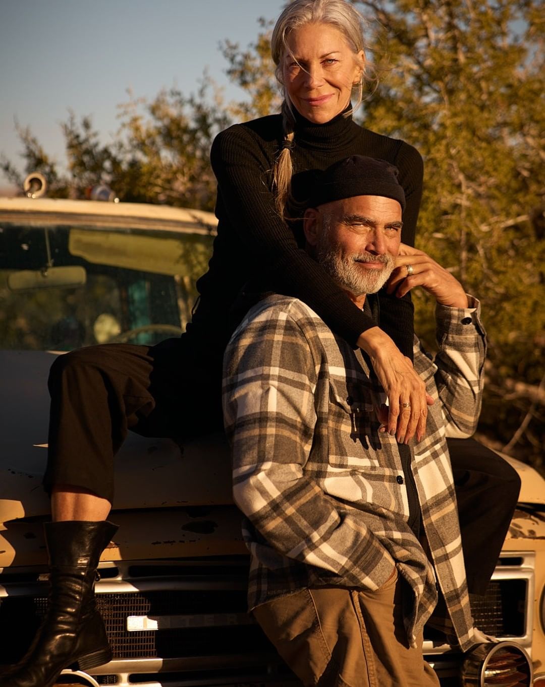 A couple poses together outdoors, with a woman sitting on the hood of a vintage truck while resting her hands on the man standing in front.