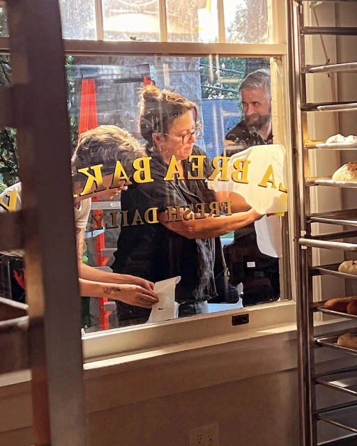 Two people assist a woman in affixing 'LA BREA BAKERY FRESH DAILY' signage to a glass window, with baked goods visible inside.