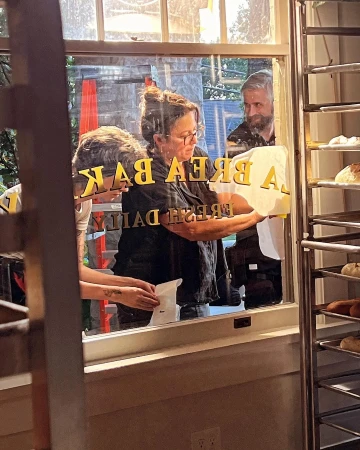 Two people assist a woman in affixing 'LA BREA BAKERY FRESH DAILY' signage to a glass window, with baked goods visible inside.