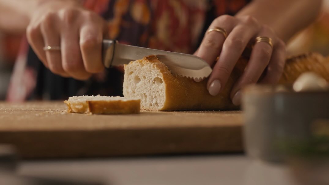 Close-up of hands slicing a baguette on a wooden board with a serrated knife. Food preparation in an indoor environment.
