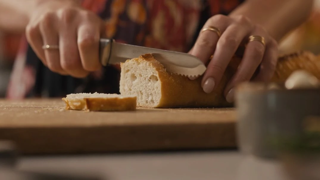 Close-up of hands slicing a baguette on a wooden board with a serrated knife. Food preparation in an indoor environment.
