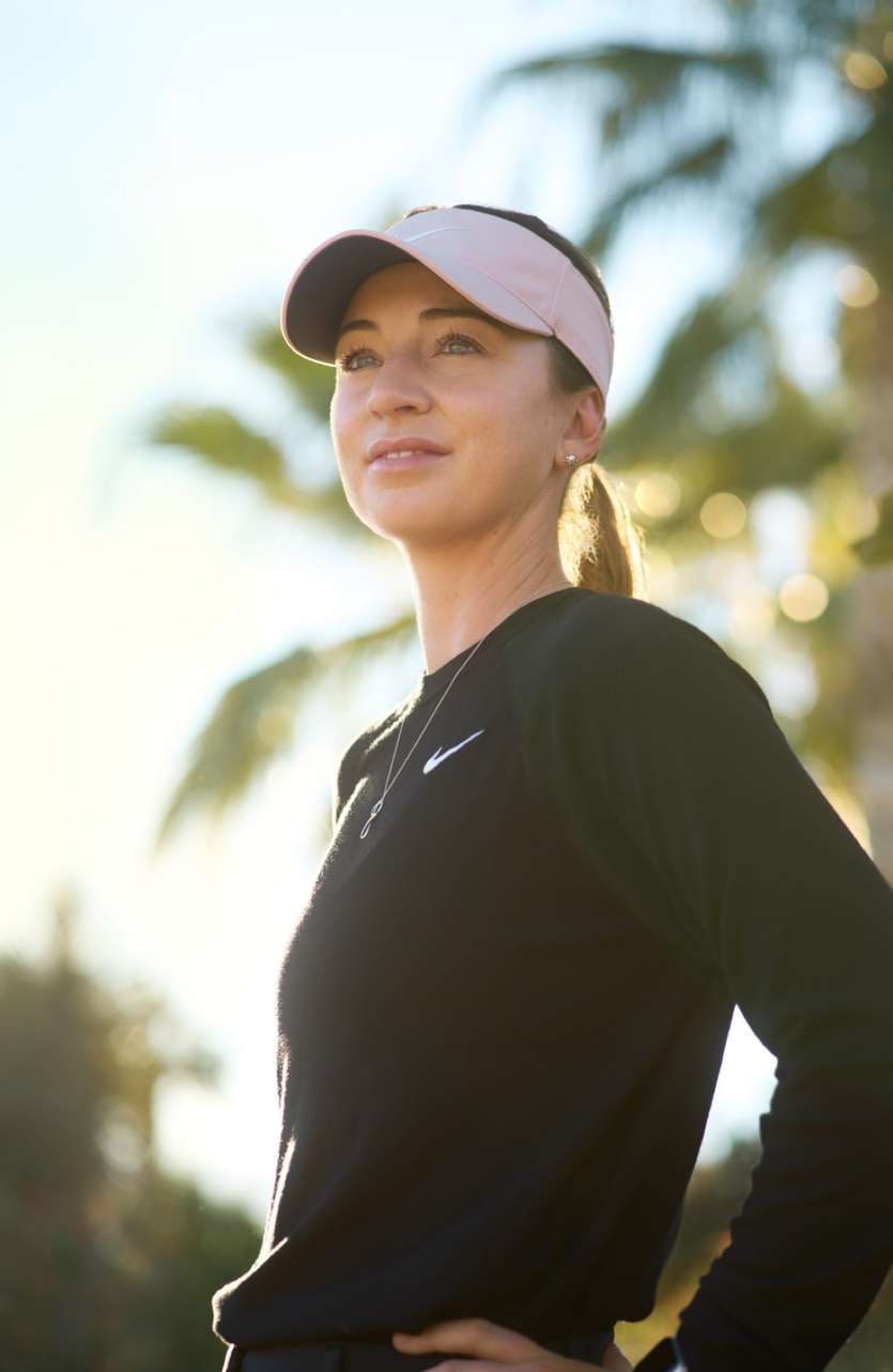 Woman wearing a black long-sleeve athletic top and a pink visor outdoors against a sunlit palm tree background.