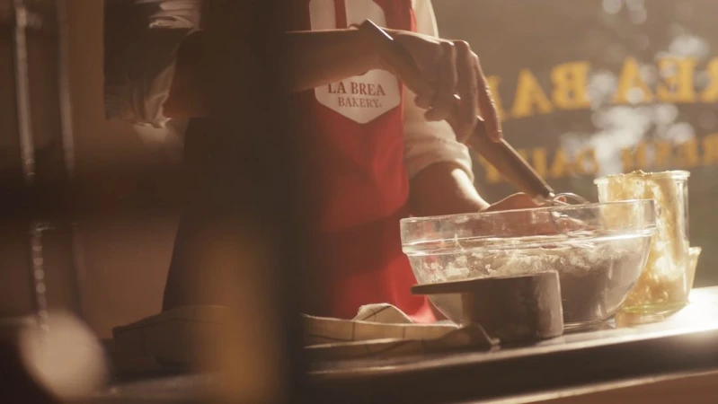 Woman in red apron stirring ingredients in clear bowl on a metal counter with bakery signage visible in the background.