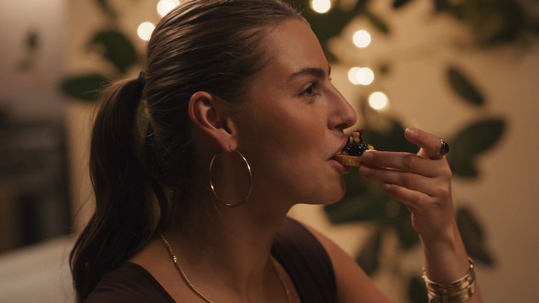 Young woman in profile eating a small tart. She wears hoop earrings, a gold chain, and has dark hair in a ponytail in soft lighting.