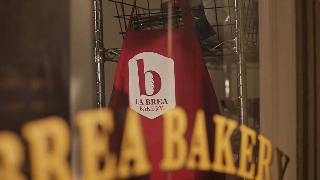 La Brea Bakery branded apron hanging in the window of a bakery, with out-of-focus text and reflections visible on the window.
