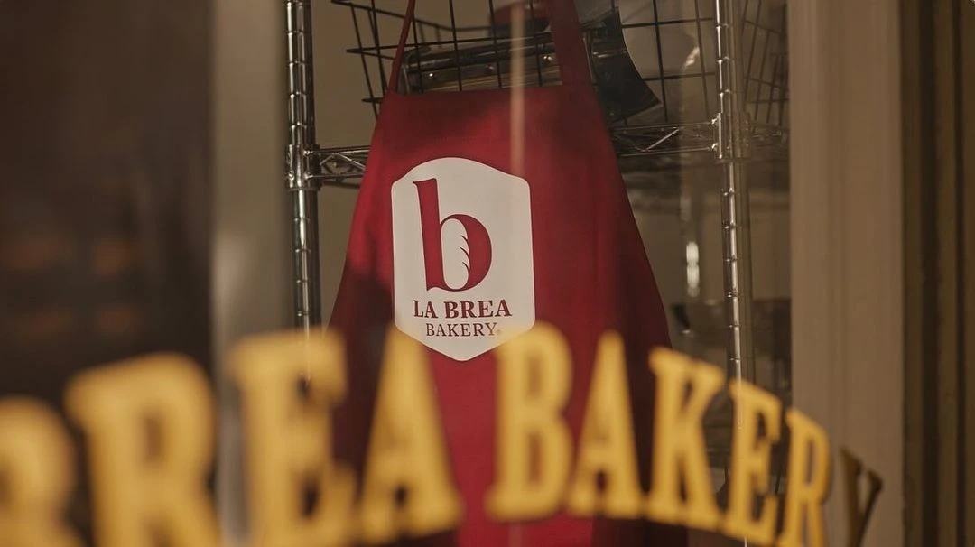 La Brea Bakery branded apron hanging in the window of a bakery, with out-of-focus text and reflections visible on the window.