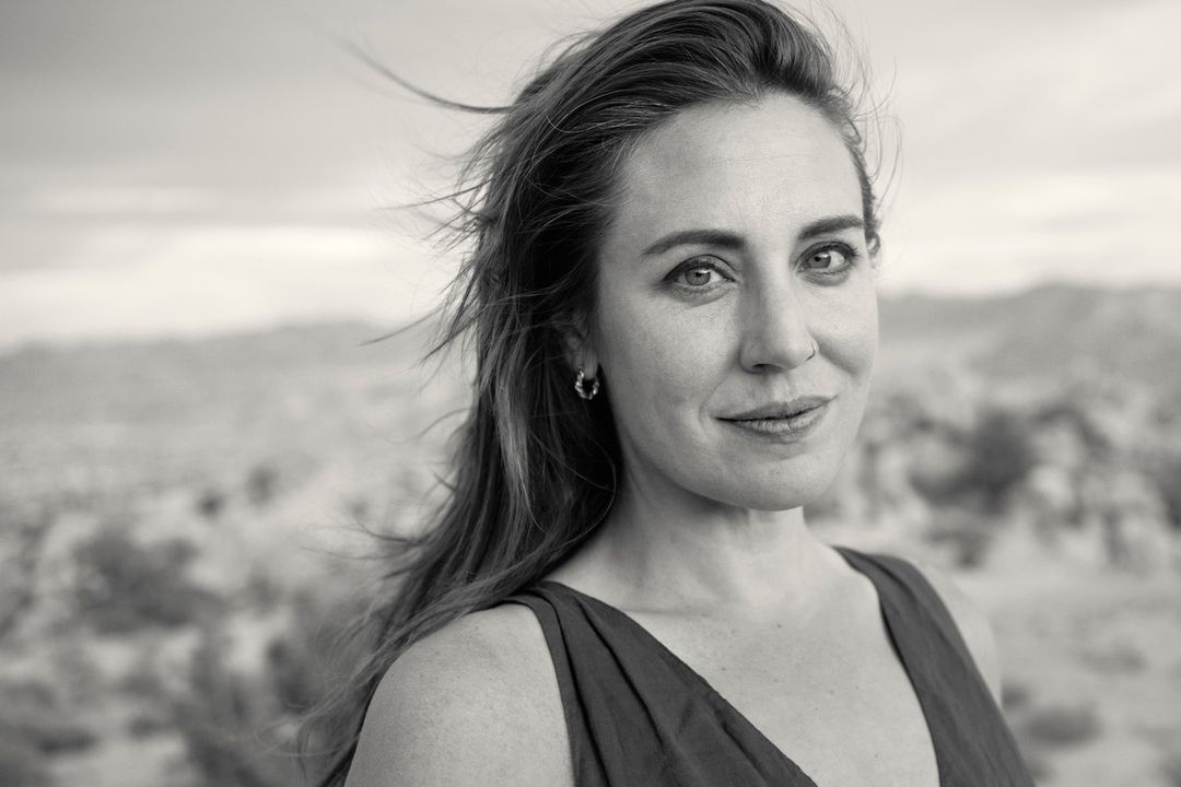 Monochrome portrait of an adult woman with hair blowing in wind, against a blurred background of rocks and sky; medium close-up shot.