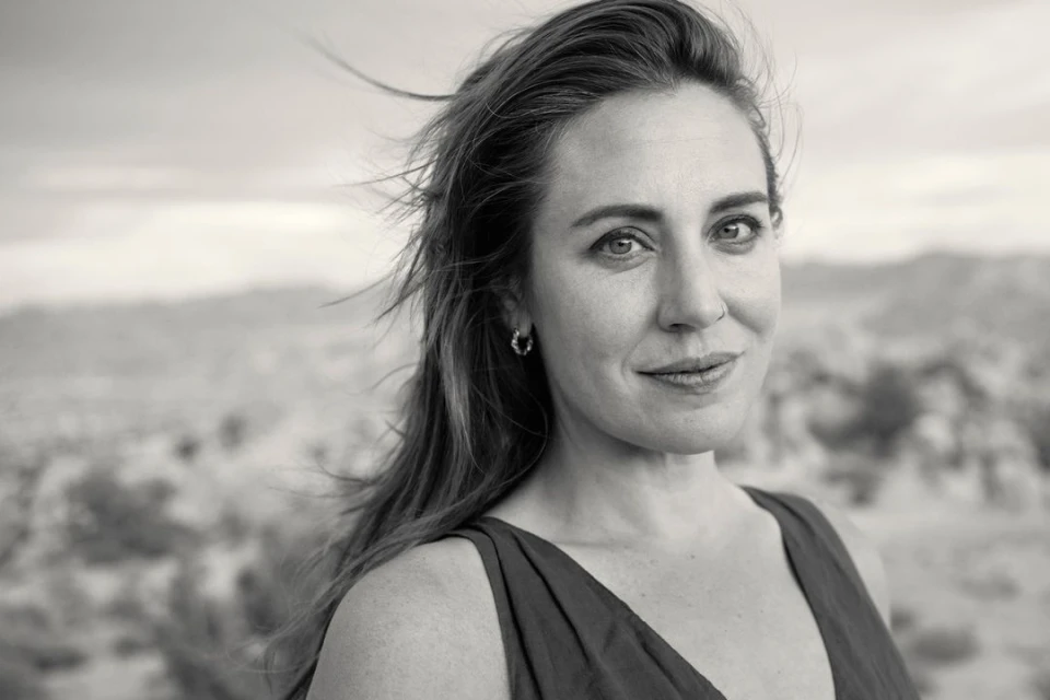 Monochrome portrait of an adult woman with hair blowing in wind, against a blurred background of rocks and sky; medium close-up shot.
