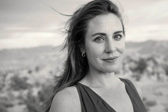 Monochrome portrait of an adult woman with hair blowing in wind, against a blurred background of rocks and sky; medium close-up shot.