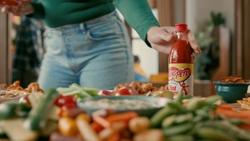Close-up of a table filled with vegetables, snack food, and a bottle of Texas Pete hot sauce, viewed from a low angle.