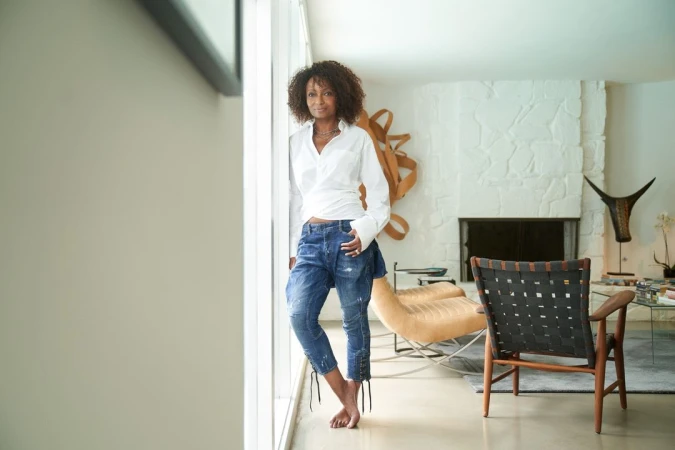 Full-length shot of an African-American woman in a white blouse and jeans standing barefoot in a brightly lit modern living room.