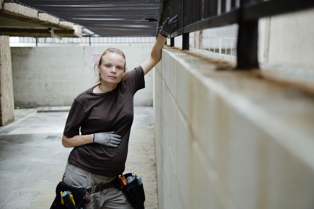 Woman in brown t-shirt with a tool belt standing in an urban environment. The photograph is taken at eye-level with medium framing.