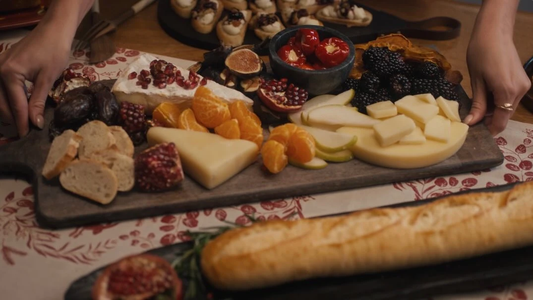 Gourmet charcuterie board featuring assorted cheeses, fruits, breads, and peppers arranged on a wooden serving board with warm lighting.