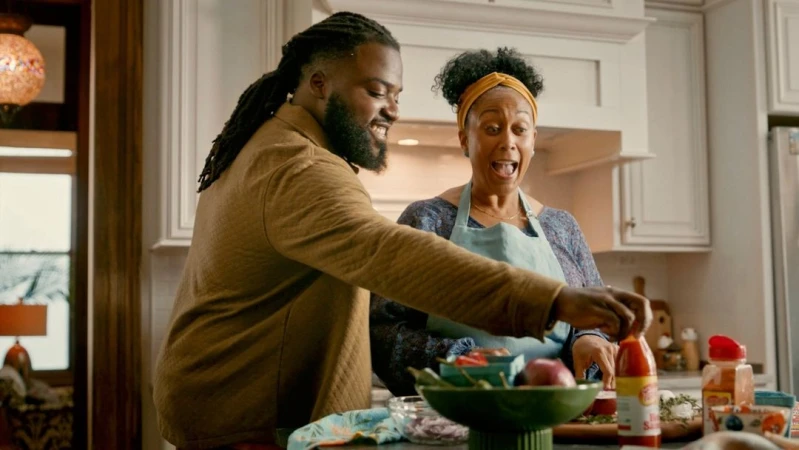 An adult male and adult female cooking together in a brightly lit kitchen, with the woman reacting in surprise or excitement to an ingredient.