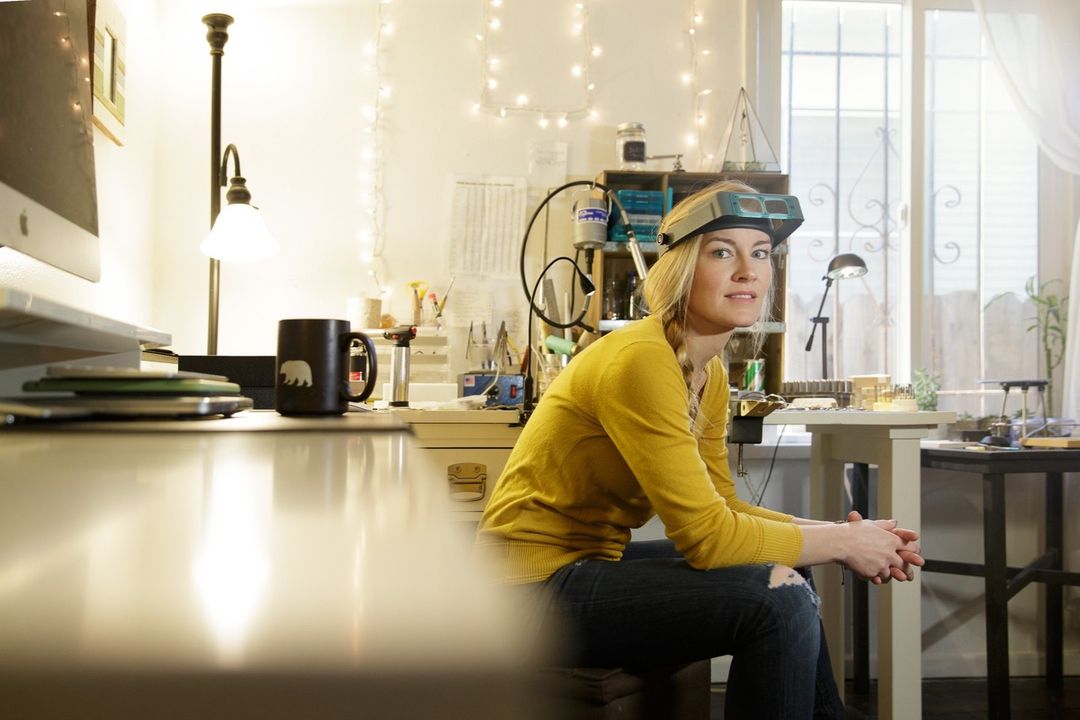A blonde adult female sits in her light-filled workshop wearing a head-mounted magnifier, with jewelry-making tools and supplies visible.