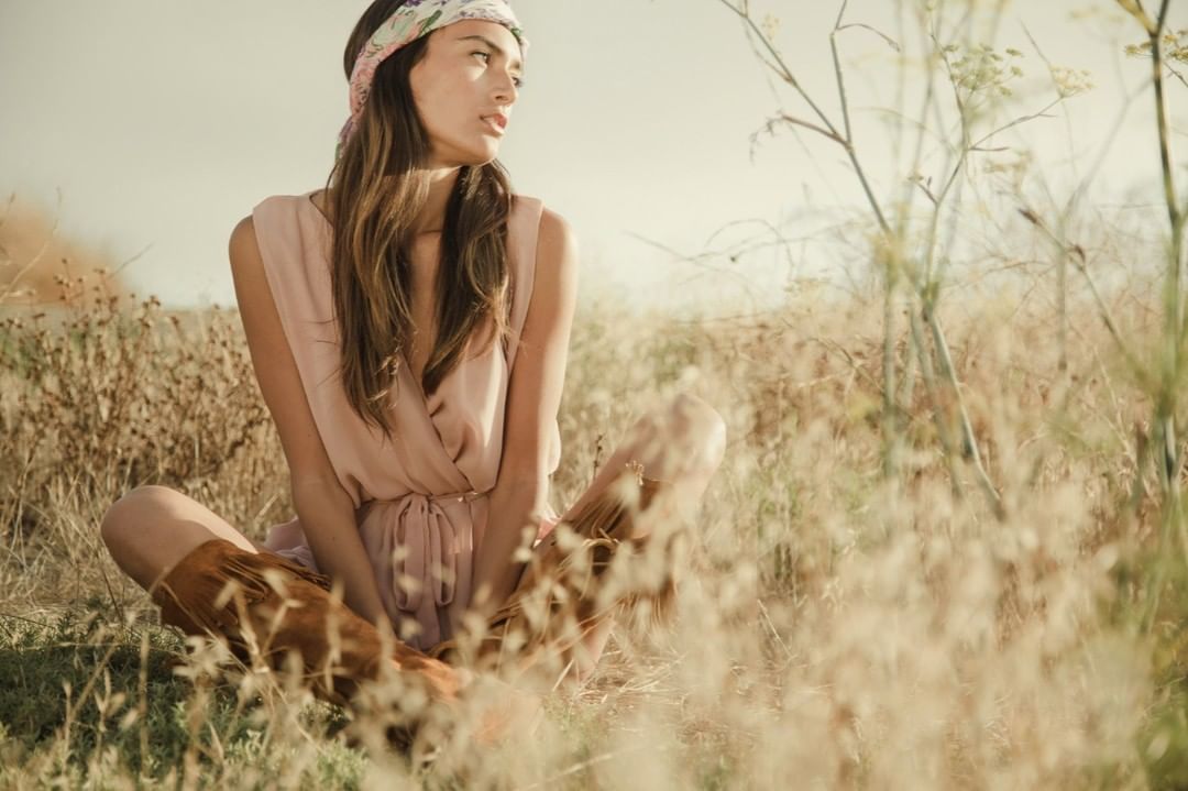 Woman in a floral headband and blush dress sits cross-legged in a grassy field with tall grass, illuminated by soft, natural light. 