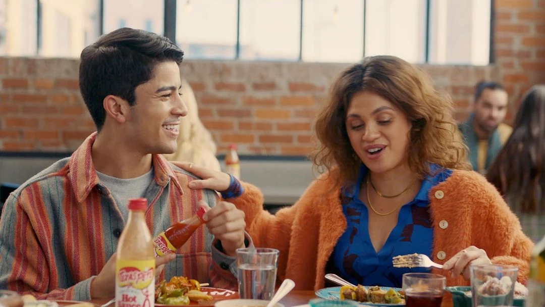 Young couple enjoys lunch together at a restaurant, sharing food and hot sauce, with other diners visible in the background.