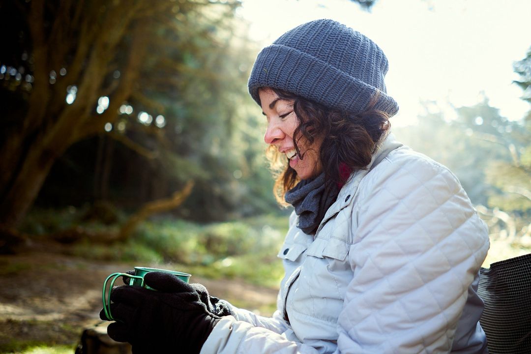 Woman wearing a beanie and quilted jacket smiles as she sits outside, holding a steaming mug in a forest setting in soft light.