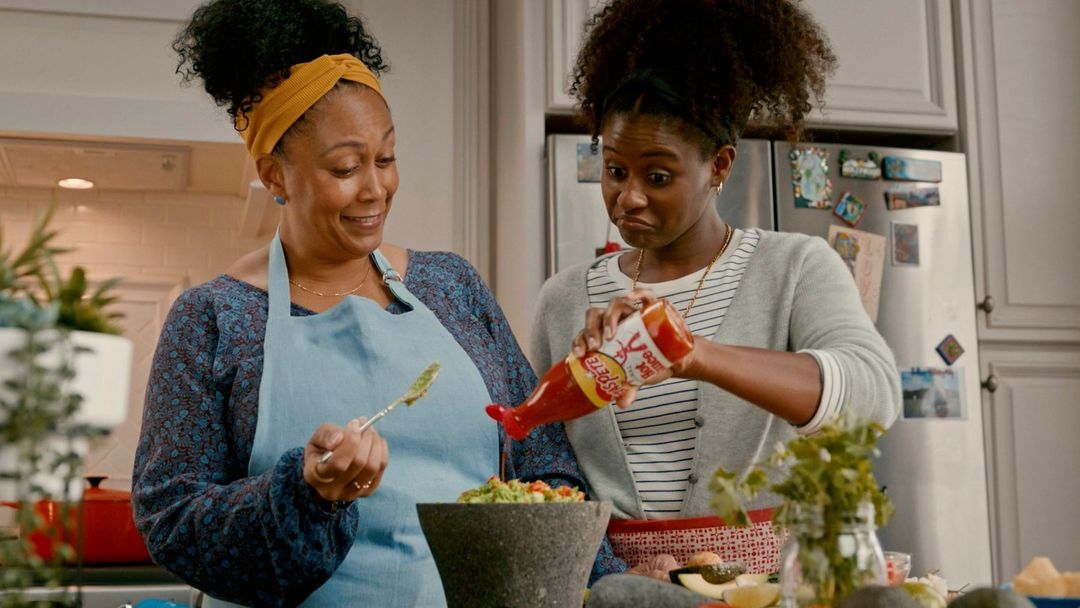 Two women in a kitchen preparing food, one adding hot sauce to guacamole. Kitchen has white cabinets and stainless steel refrigerator.