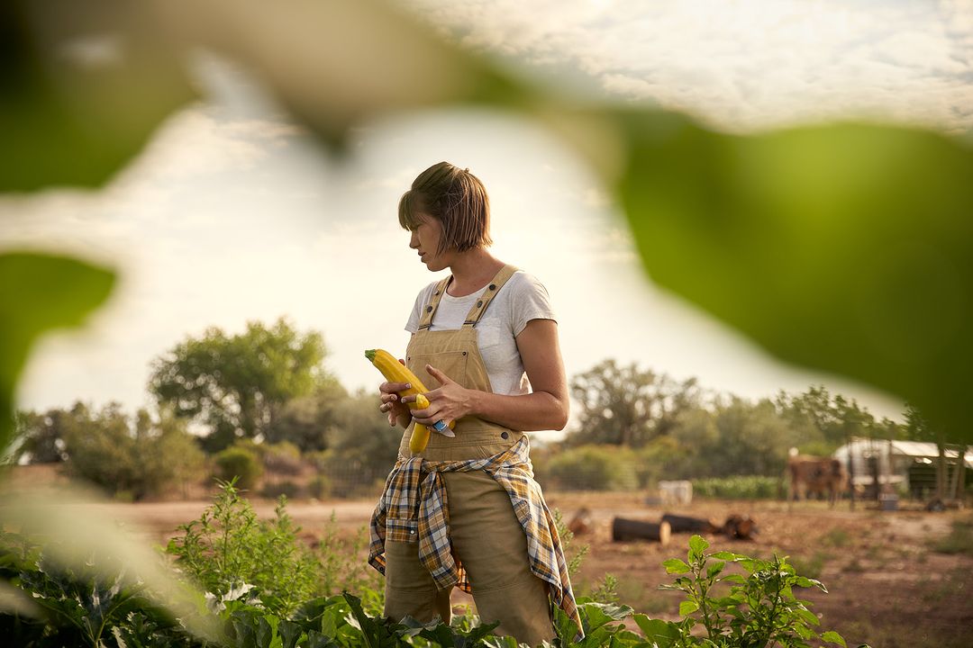 Woman in overalls holds fresh yellow squash in an outdoor farm setting with lush green plants and trees against a cloudy sky.