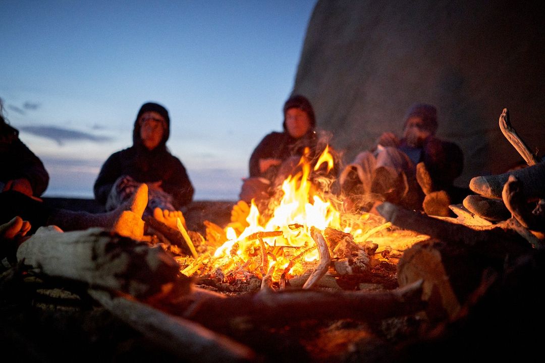 People gathered around a bonfire at dusk with dark log pile. A group of people are enjoying the firelight during the evening.