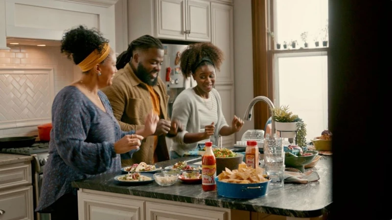 Three people smiling and dancing in a modern domestic kitchen around a marble-topped island with plates of food.