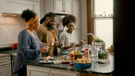 Three people smiling and dancing in a modern domestic kitchen around a marble-topped island with plates of food.