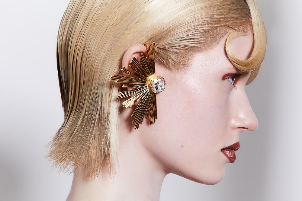 Profile portrait of a blonde woman with slicked-back hair and an ornate gold ear cuff, set against a plain, light background in studio lighting.