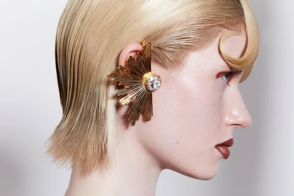 Profile portrait of a blonde woman with slicked-back hair and an ornate gold ear cuff, set against a plain, light background in studio lighting.