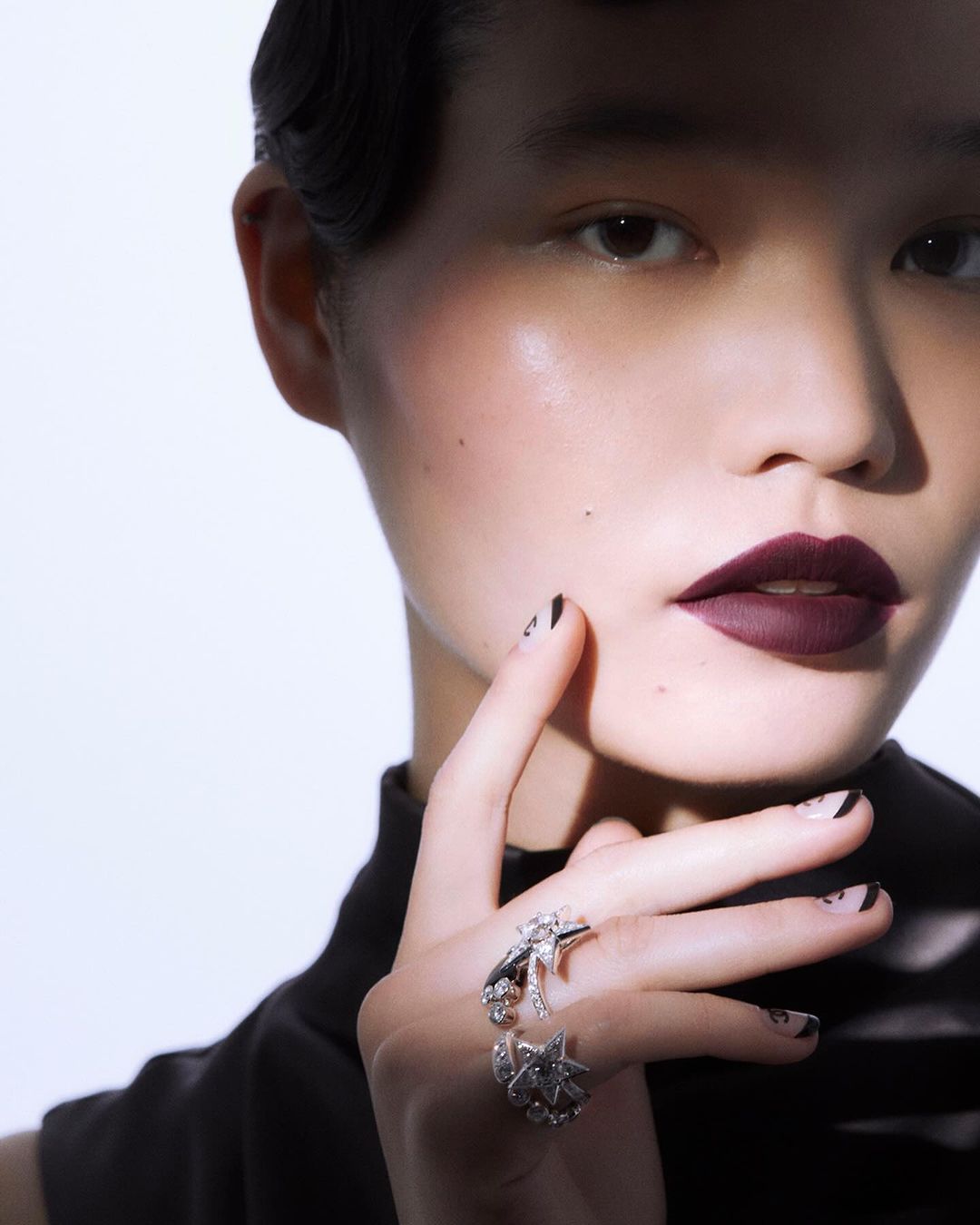 Close-up of a woman with dark lipstick and patterned nails wearing an animal-themed ring, posing against a bright white background.