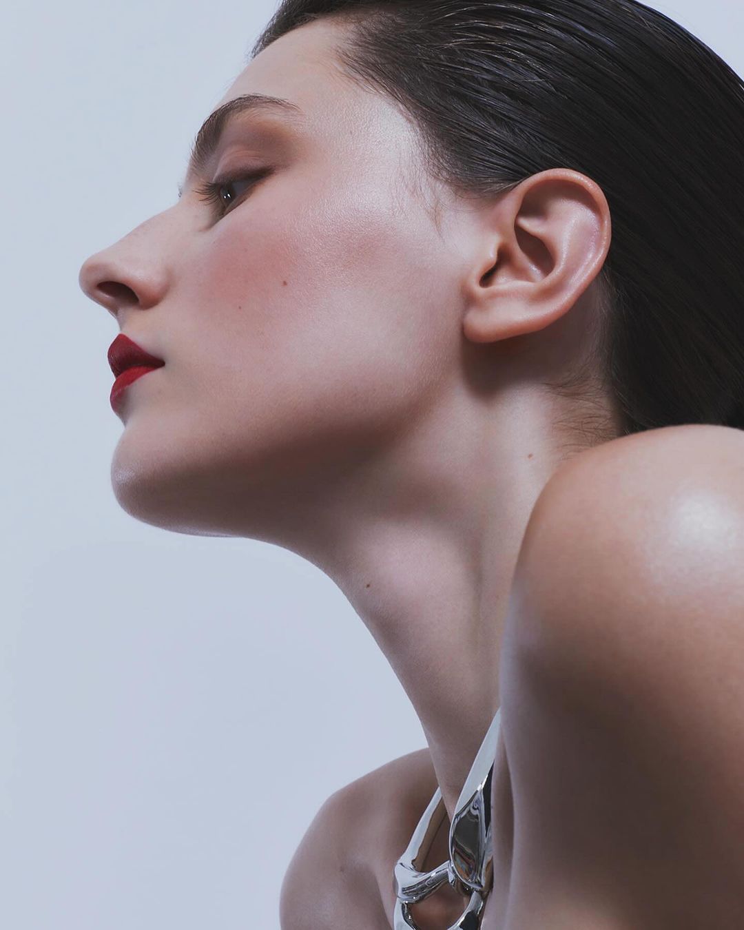 Close-up profile of a woman with dark hair, red lipstick, and a metallic top against a plain background in a beauty or fashion context.