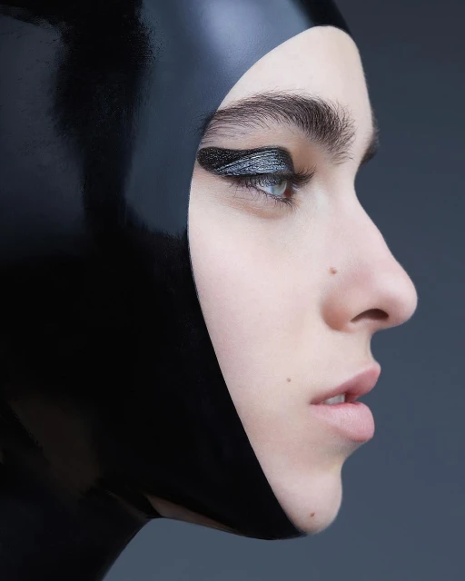 Close-up of a woman with dramatic silver and black eye makeup wearing a sleek black hood against a dark grey background.