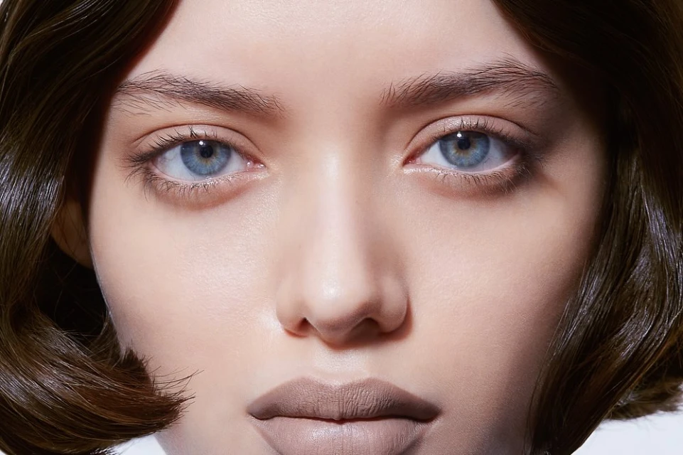 Close-up portrait of a woman's face, highlighting her blue eyes, fair skin, and nude lipstick, set against a neutral background.