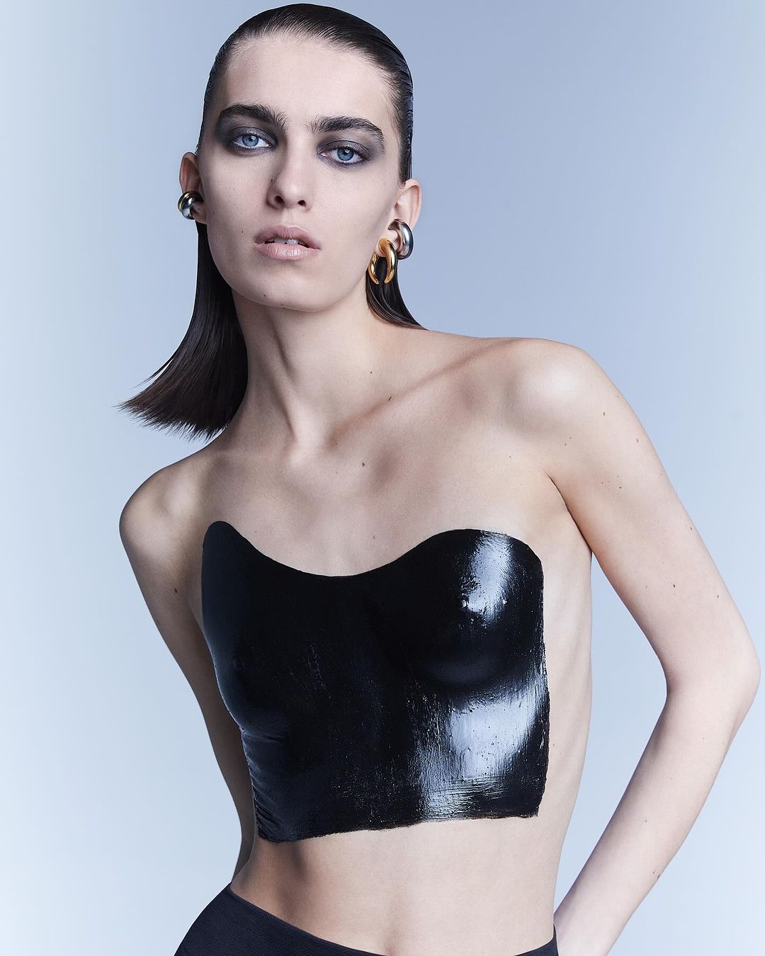 Close-up shot of a model with slicked-back hair and black body paint on a light grey background, showcasing makeup and body art.