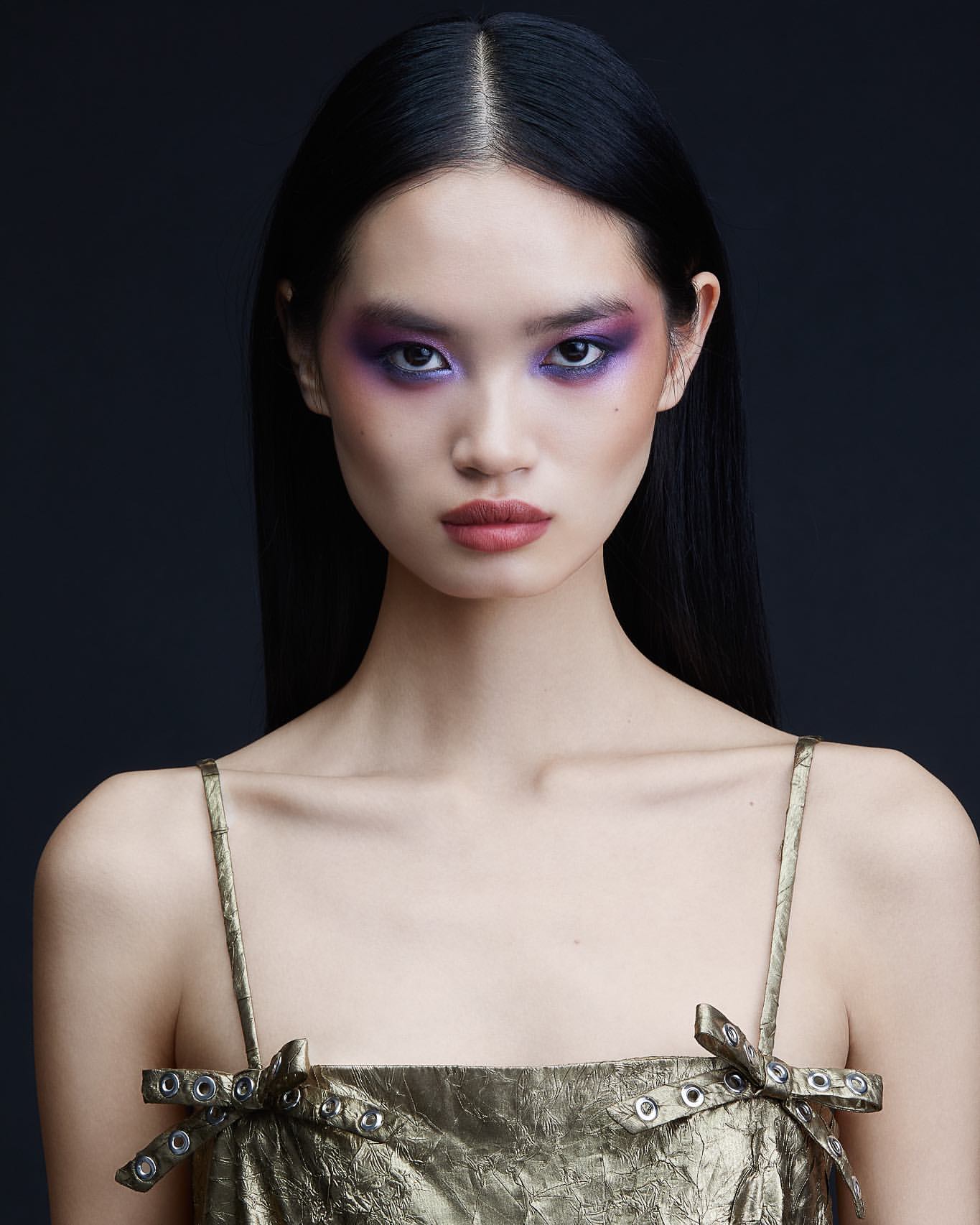 Portrait of an East Asian woman with long black hair, purple eye makeup, and metallic gold dress against a dark background by Natasja Fourie