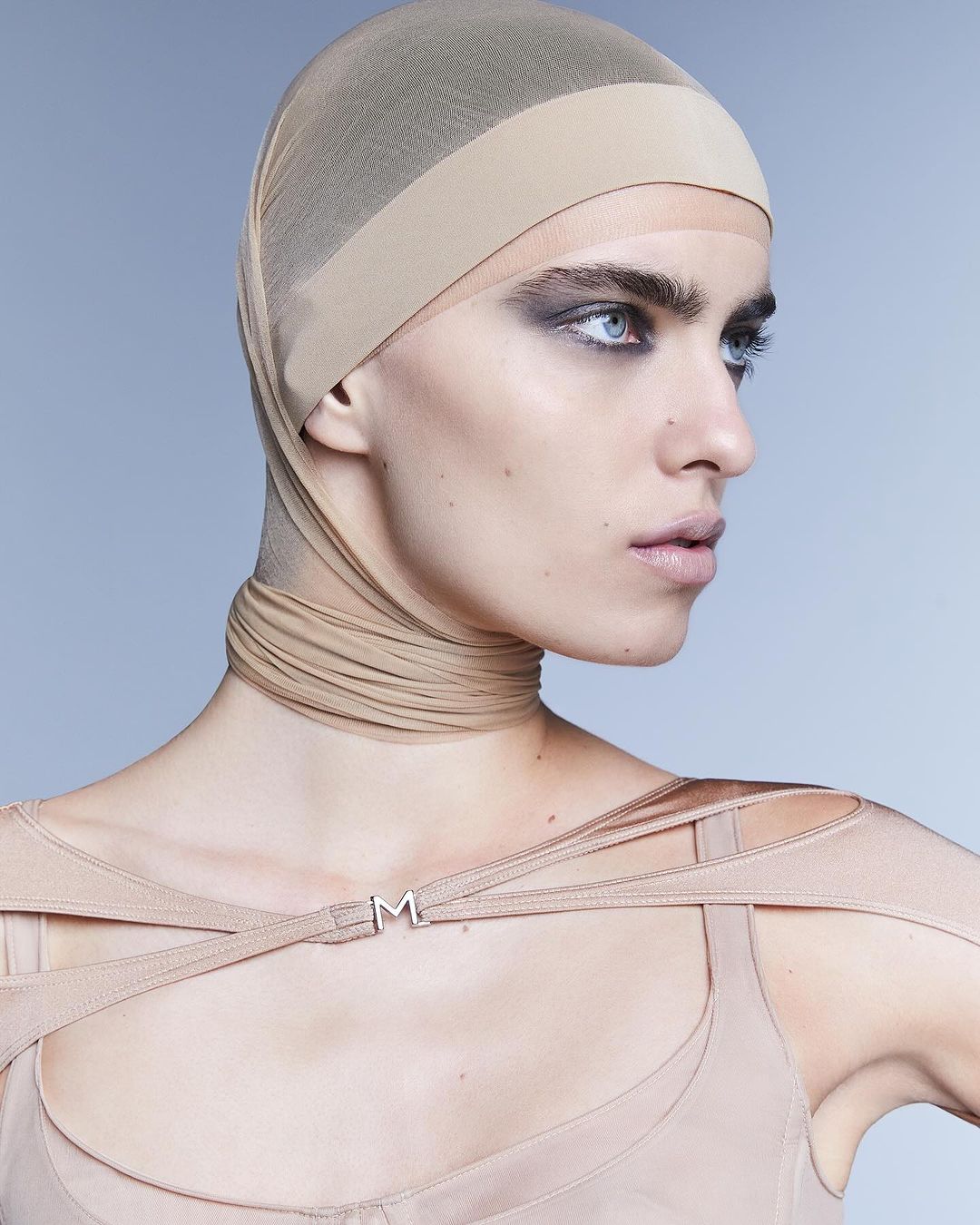 Headshot of a young adult woman with fair skin wearing a beige head covering, smoky eye makeup, and a beige top against a light blue background.