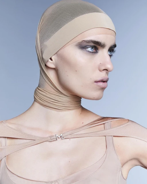 Headshot of a young adult woman with fair skin wearing a beige head covering, smoky eye makeup, and a beige top against a light blue background.