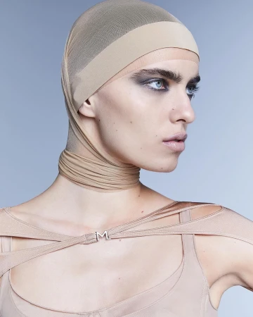 Headshot of a young adult woman with fair skin wearing a beige head covering, smoky eye makeup, and a beige top against a light blue background.